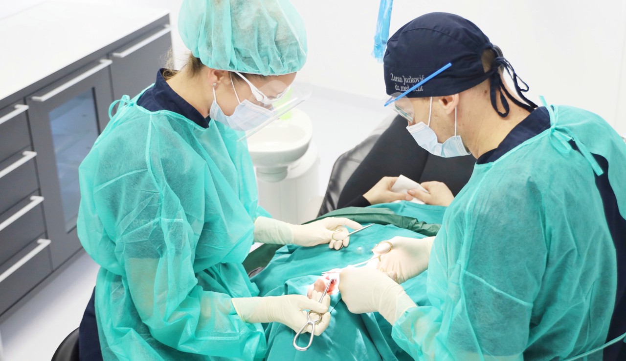 Doktor i asistent u kirurškoj opremi fokusirano izvode stomatološki zahvat u sterilnim uvjetima ordinacije.