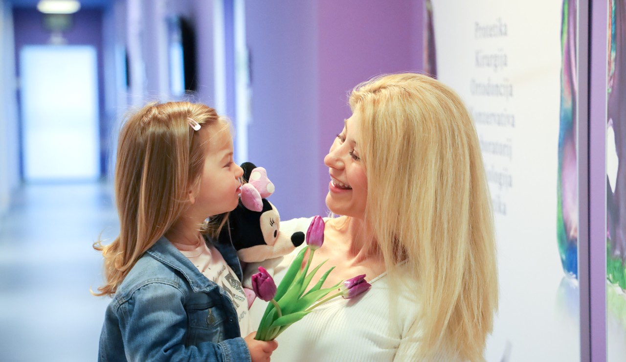 Majka s osmijehom drži dijete u naručju dok stoje u hodniku dentalne klinike, stvarajući toplu i sigurnu atmosferu.