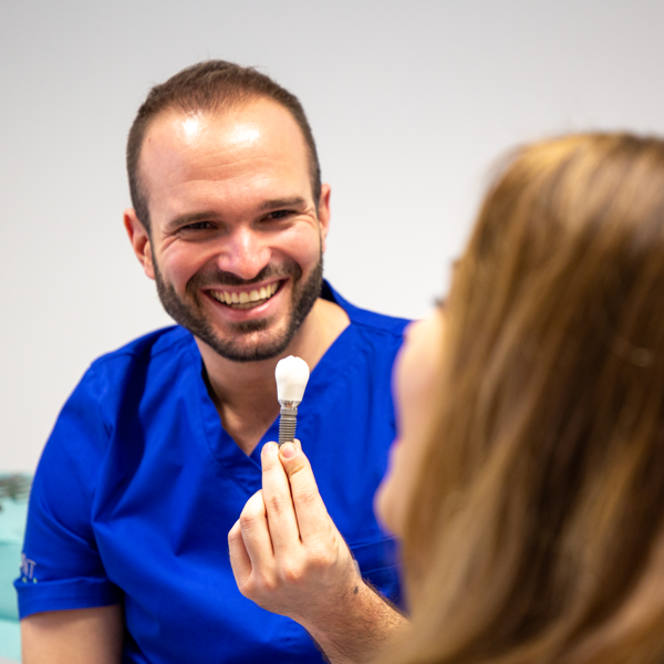 Doktor s osmijehom drži u ruci model zubnog implantata i objašnjava pacijentici kako funkcionira terapija implantatima.