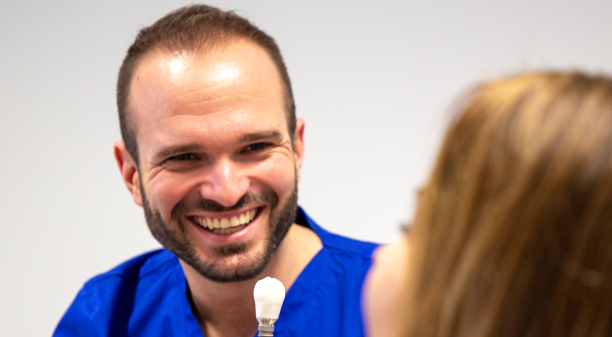 Doktor s osmijehom drži u ruci model zubnog implantata i objašnjava pacijentici kako funkcionira terapija implantatima.