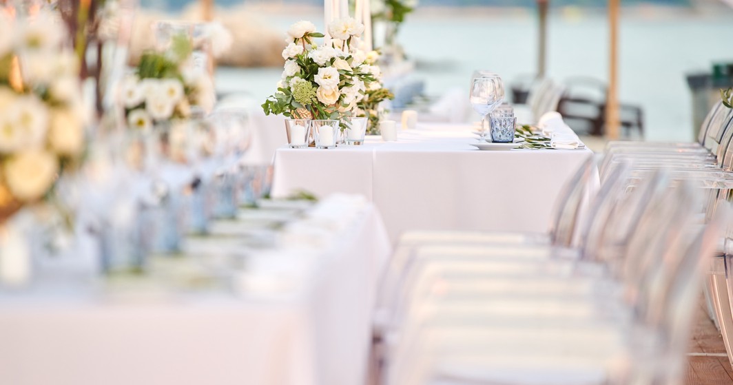 L'immagine mostra un allestimento per matrimoni con dettagli di fiori e candele all'interno di una tenda, sedie trasparenti e il mare sullo sfondo.