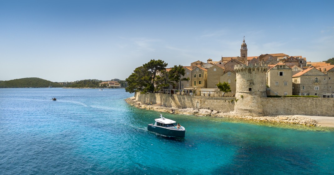 Scenski pogled na povijesni obalni grad Korčula, Hrvatska, s malim brodom koji plovi kristalno čistim Jadranskim morem u blizini Aminess Younique Korčula Heritage Hotela.