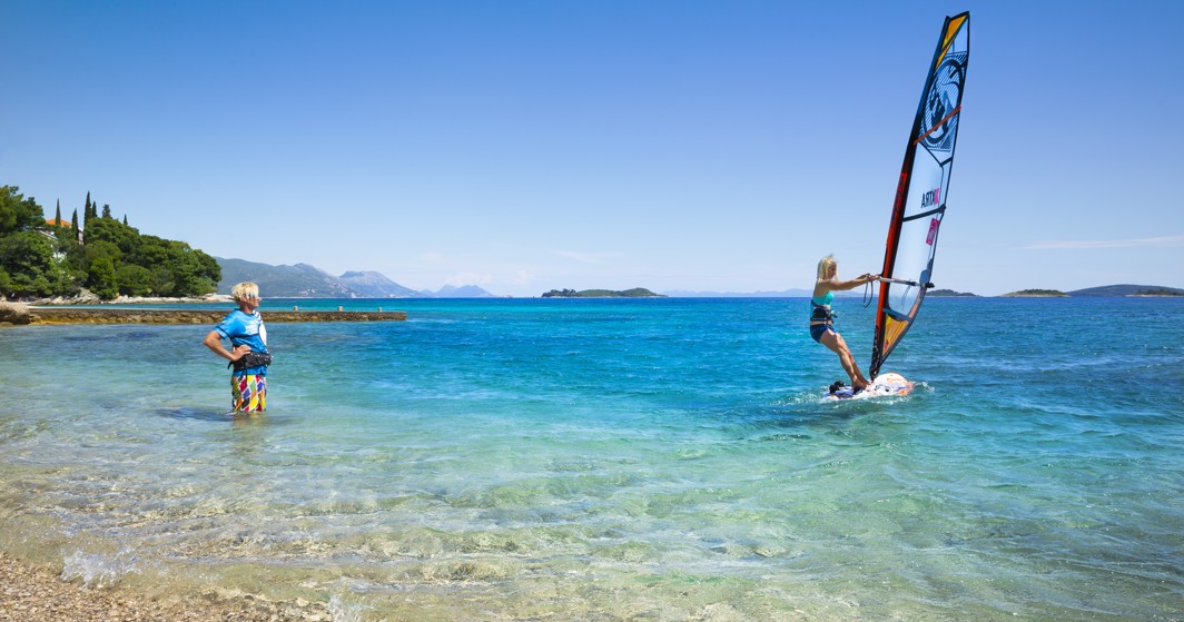 Dijete sudjeluje u surferskoj klasi u plitkim vodama Korčule, pod vodstvom instruktora, s živopisnim, sunčanim morskim krajolikom blizu Maradiso Marko Polo by Aminess.