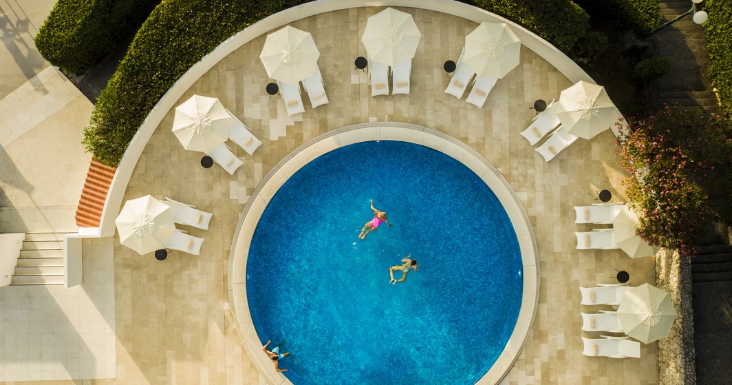 Una vista aerea di una piscina con persone che nuotano, caratterizzata da lettini per prendere il sole e rilassarsi sotto gli ombrelloni presso l'Aminess Younique Liburna Hotel.