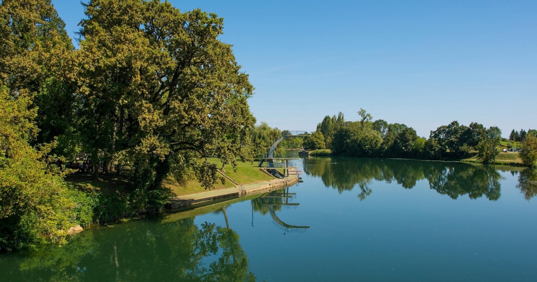 Area balneare sul fiume con parco, alberi e ponte riflesso nell’acqua calma