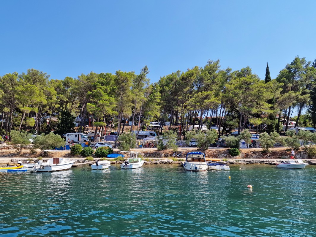 Mirno obmorsko kampiranje v Aminess Port9 Planet Camping Korcula, z šotori in avtodomi v senci bujnih borovcev, s čistim pogledom na mirno turkizno vodo in naravno obalo