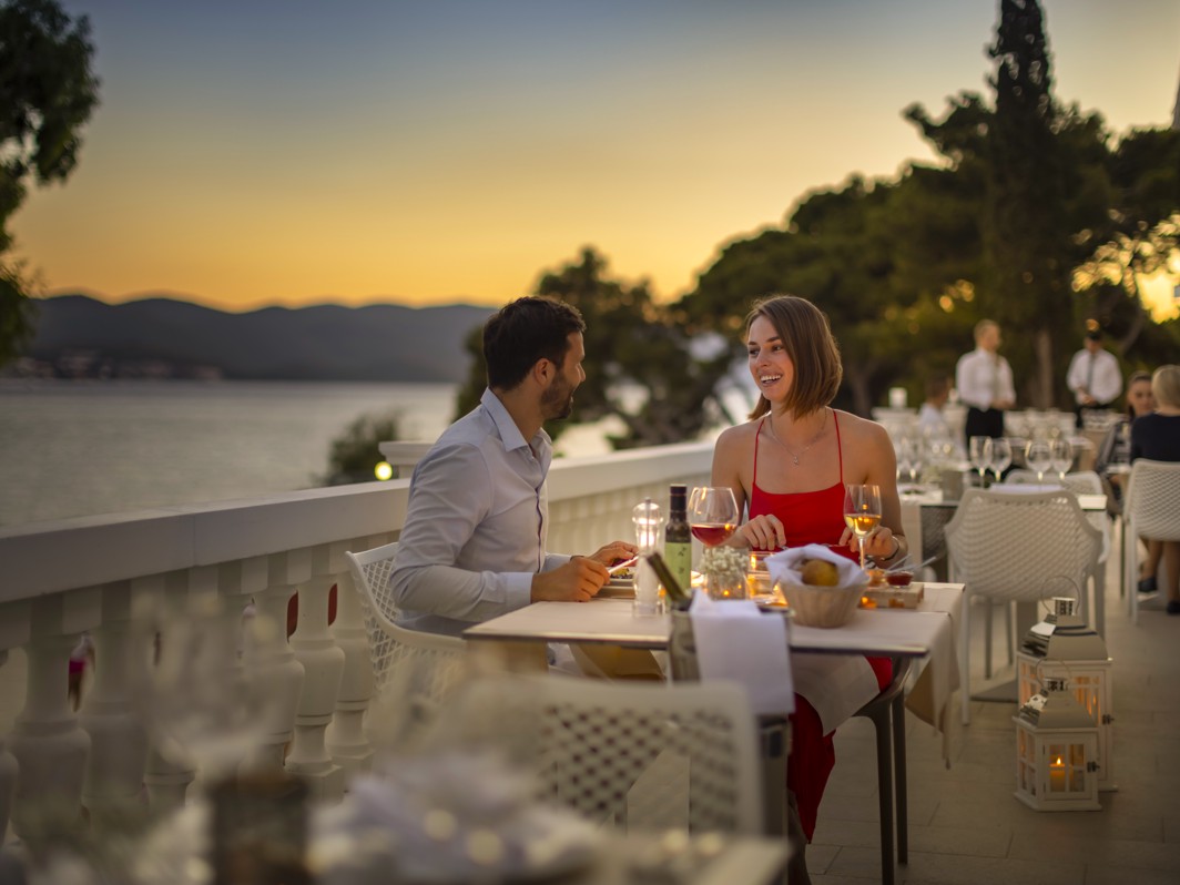 A couple dining at the terrace of a restaurant, overlooking a serene sunset by the water at Aminess Maradiso Bellevue Village.