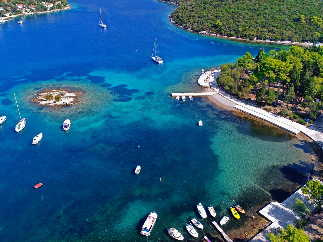 Osupljiv zračni pogled na Korčulanski zaljev blizu Aminess Port9 Planet Camping, prikazuje živahne tirkizne vode, male sidrene čamce in bujno zeleno obalo prekrito z gozdom