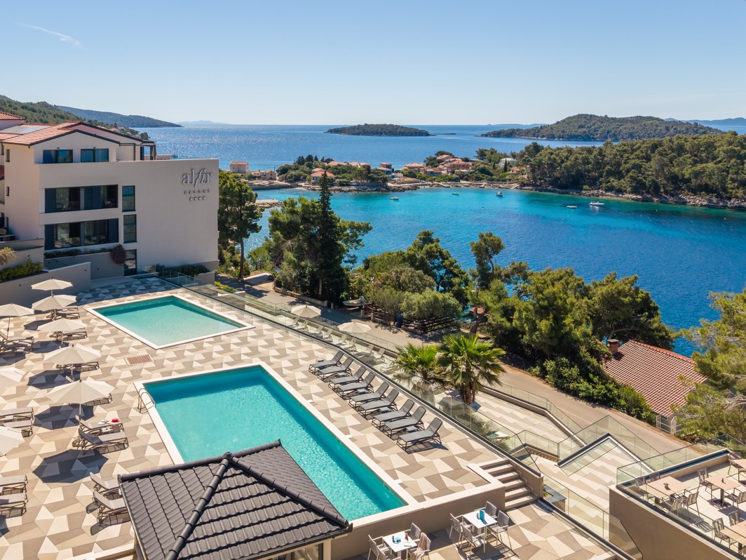 Zwembaden van Aminess Alfir Hotel met uitzicht op turquoise zee en dennenkust.