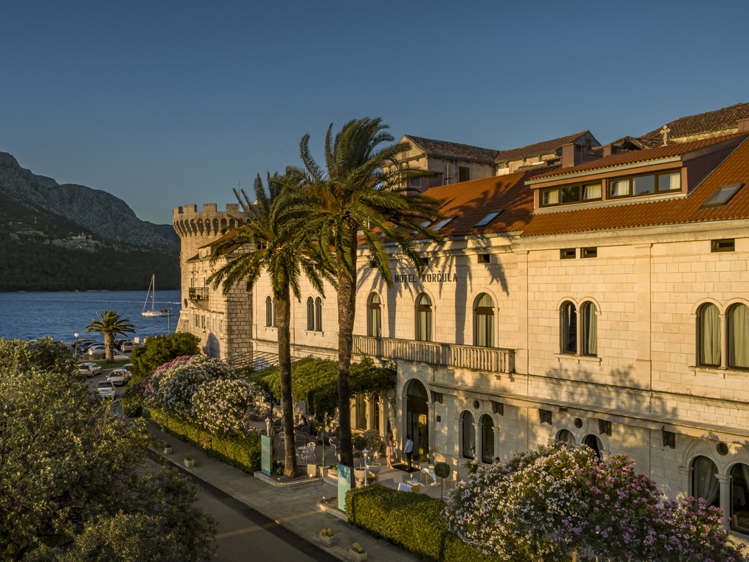 Aminess Younique Korcula Heritage Hotel, a historic building with red-tiled roofs in Korcula's Old Town near the sea, located beside Tower Kanalevic, surrounded by palm trees and blooming gardens, captured during golden hour.