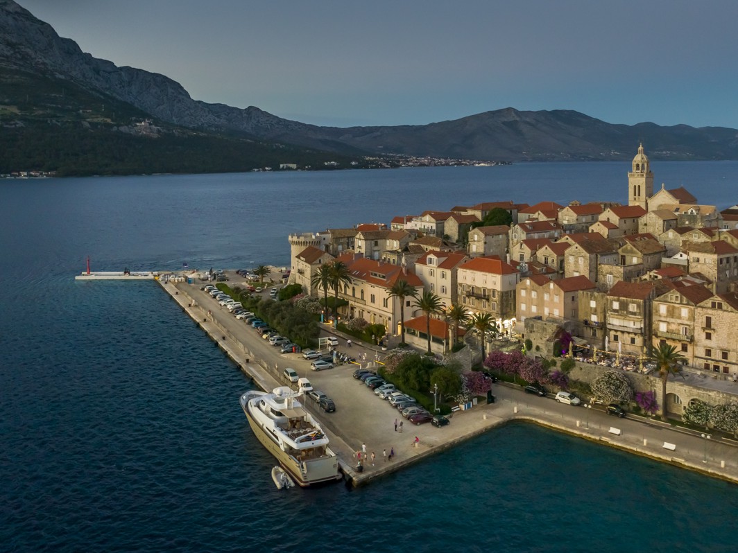 Drone view of Korcula Old Town at dusk, featuring Aminess Younique Korcula Heritage Hotel along the promenade near the port, with red-tiled rooftops, palm trees, and the Peljesac Channel in the background.