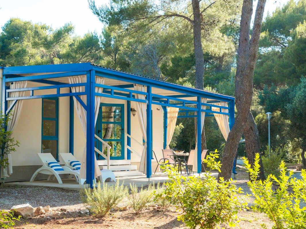 Pogled izvana na Marko Polo Holiday Home u Port9 Planet Camping by Aminess, Korčula, sa strukturama uokvirenim plavim bojama okruženim borovima pod vedrim nebom.