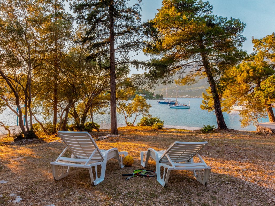 Slikovit kamp prostor s pogledom na more u Aminess Port9 Planet Camping, Korčula, s ležaljkama postavljenim pod visokim drvećem, s pogledom na vodu i okružen šumovitim krajolikom