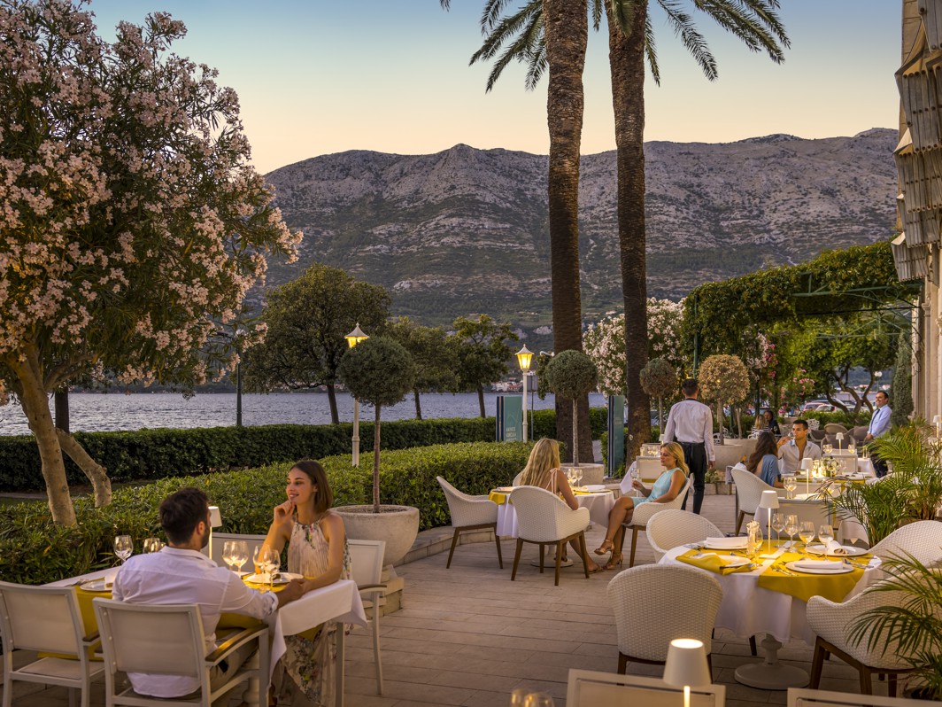 Guests enjoying a romantic dinner on the terrace of 7 Seas Restaurant & Bar at Aminess Korcula Heritage Hotel at sunset, surrounded by blooming oleander trees, palm trees, and a view of the Peljesac hills across the sparkling sea.