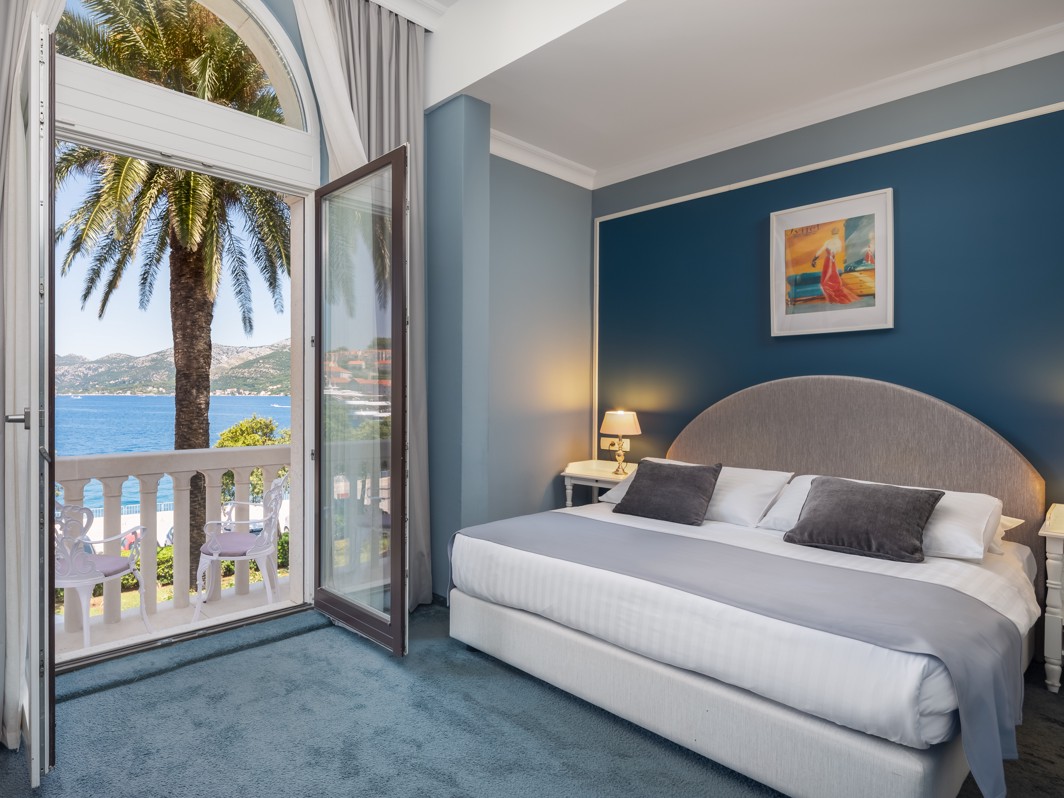 Elegant superior double room with a comfortable bed, stylish decor in blue tones, and a balcony offering stunning seaside views, framed by a palm tree, at Aminess Younique Korcula Heritage Hotel.