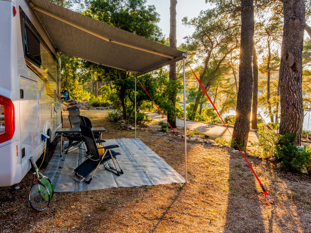 Avtokamp na lokaciji Aminess Port9 Planet Camping Korcula, kjer je parkiran avtodom pod borovci z zasenčenim prostorom za sedenje in pogledom na okoliško naravno pokrajino