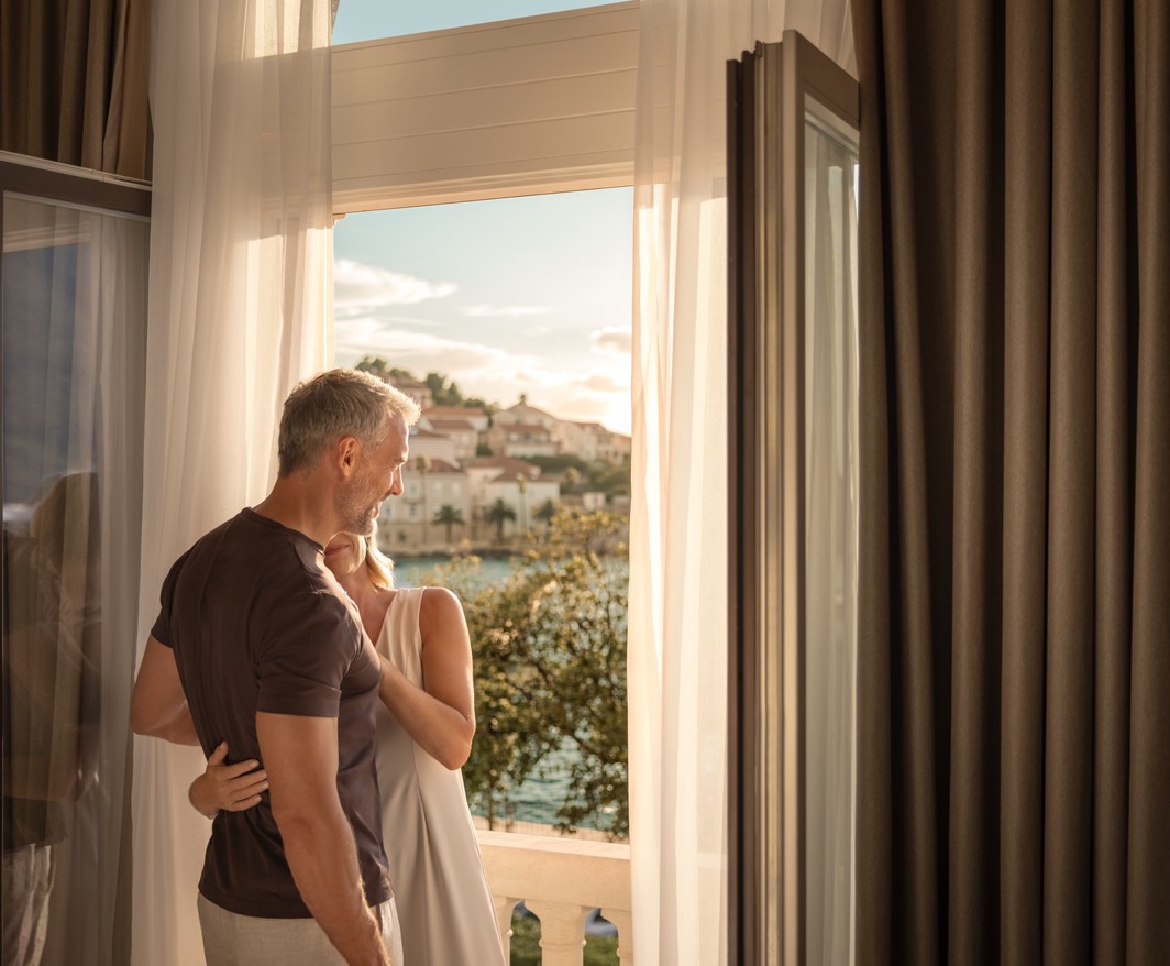 A couple embraces by the open balcony at Aminess Younique Korcula Heritage Hotel at sunset.