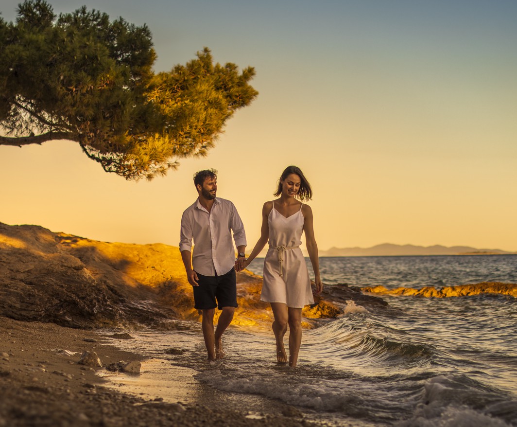 Paar spaziert bei Sonnenuntergang entlang der Küste in der Nähe von Aminess Maradiso Casa Bellevue, mit goldenen Himmeln und ruhigen Gewässern im Hintergrund.