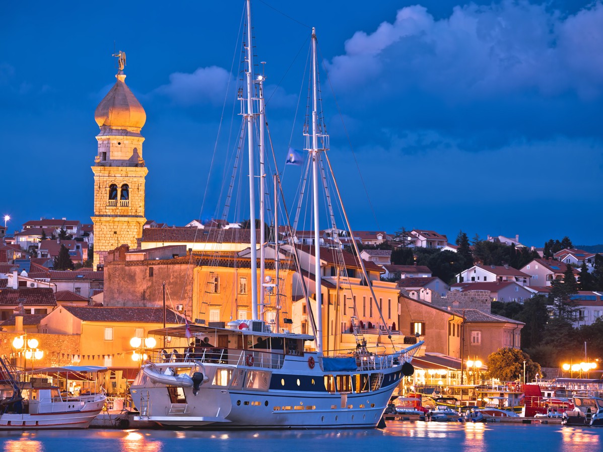 Night view of Krk harbor, Croatia, featuring illuminated sailboats and the historic bell tower.