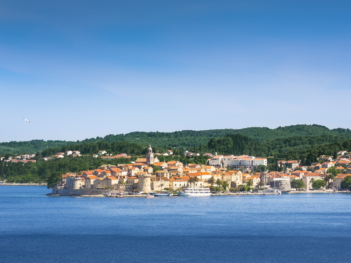 Stari grad Korčula na otoku Korčuli s terakota krovovima, zelenim brežuljcima i Jadranskim morem pod vedrim plavim nebom.