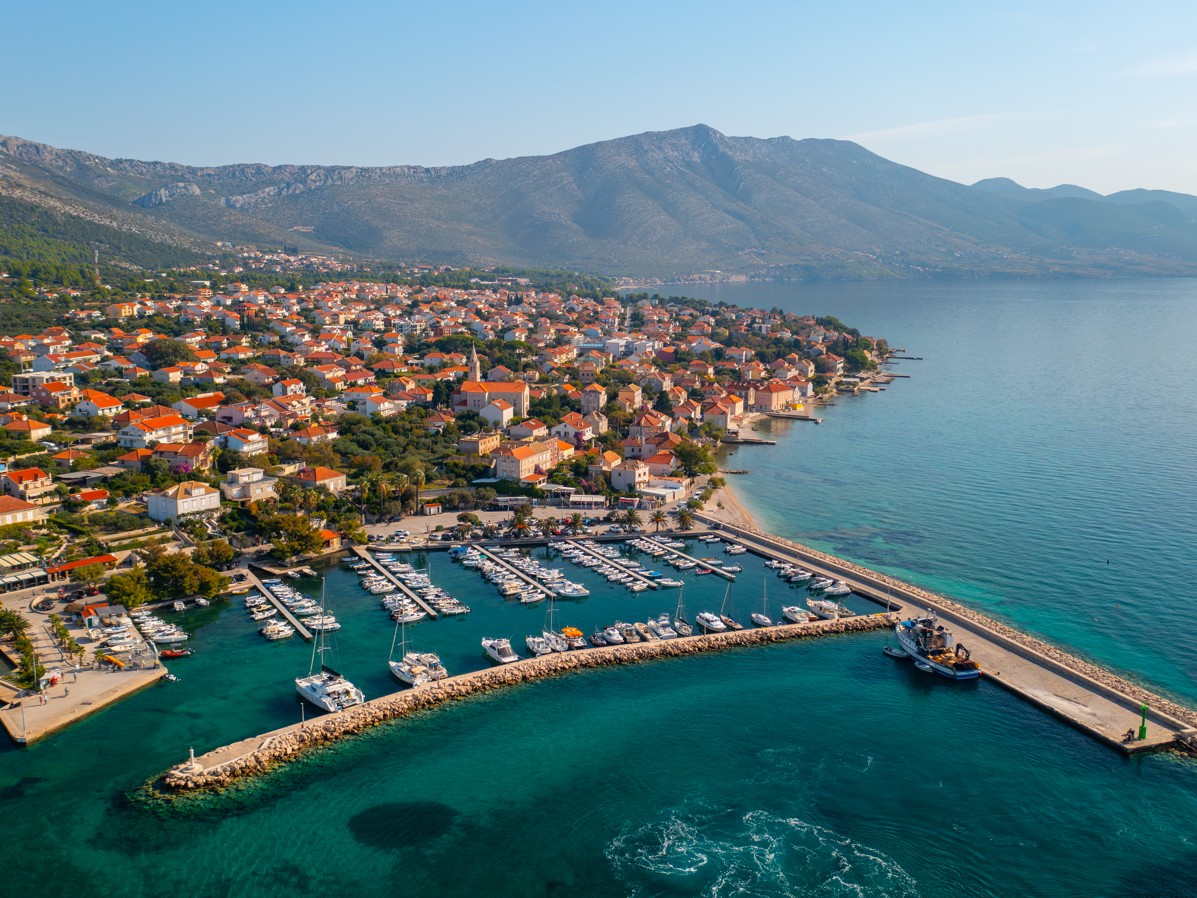 Zračni pogled na Orebić na poluotoku Pelješac, s marinom, kućama s crvenim krovovima i Jadranskim morem u pozadini planina.