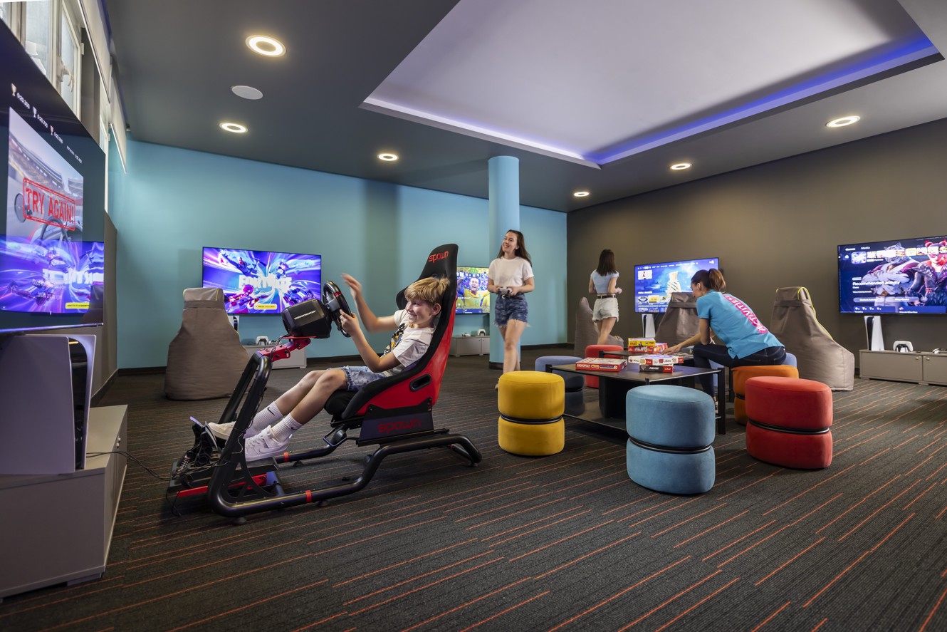 L'immagine mostra un ragazzo seduto in una sedia simulatore, con animatori che giocano un gioco con i bambini sullo sfondo.