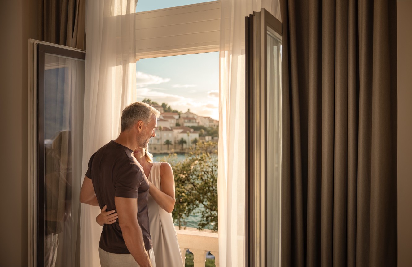 A couple embraces by the open balcony at Aminess Younique Korcula Heritage Hotel at sunset.