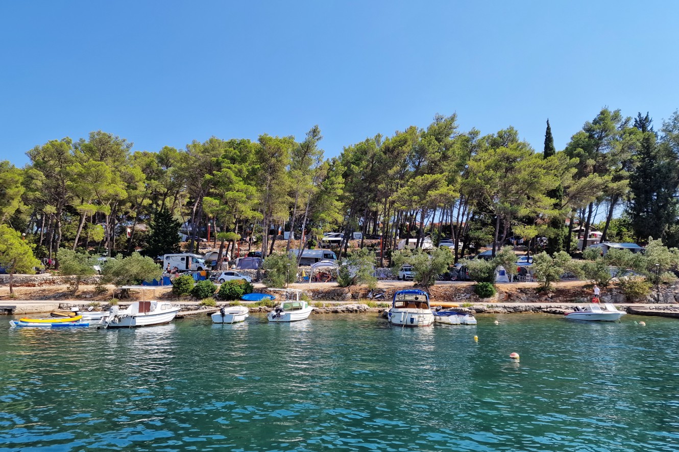 Eine ruhige Campingplatz am Meer bei Aminess Port9 Planet Camping Korcula, mit Zelten und Wohnwagen im Schatten von üppigen Kiefern, mit klarem Blick auf das ruhige türkisfarbene Wasser und die natürliche Küstenlinie