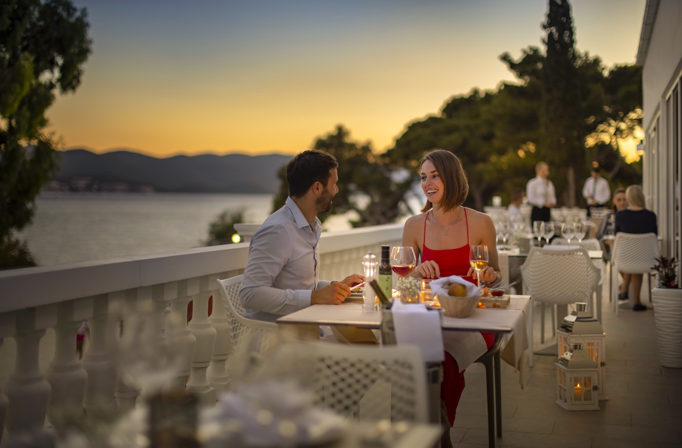 Ein Paar speist auf der Terrasse eines Restaurants und blickt auf einen ruhigen Sonnenuntergang am Wasser im Aminess Maradiso Bellevue Village.