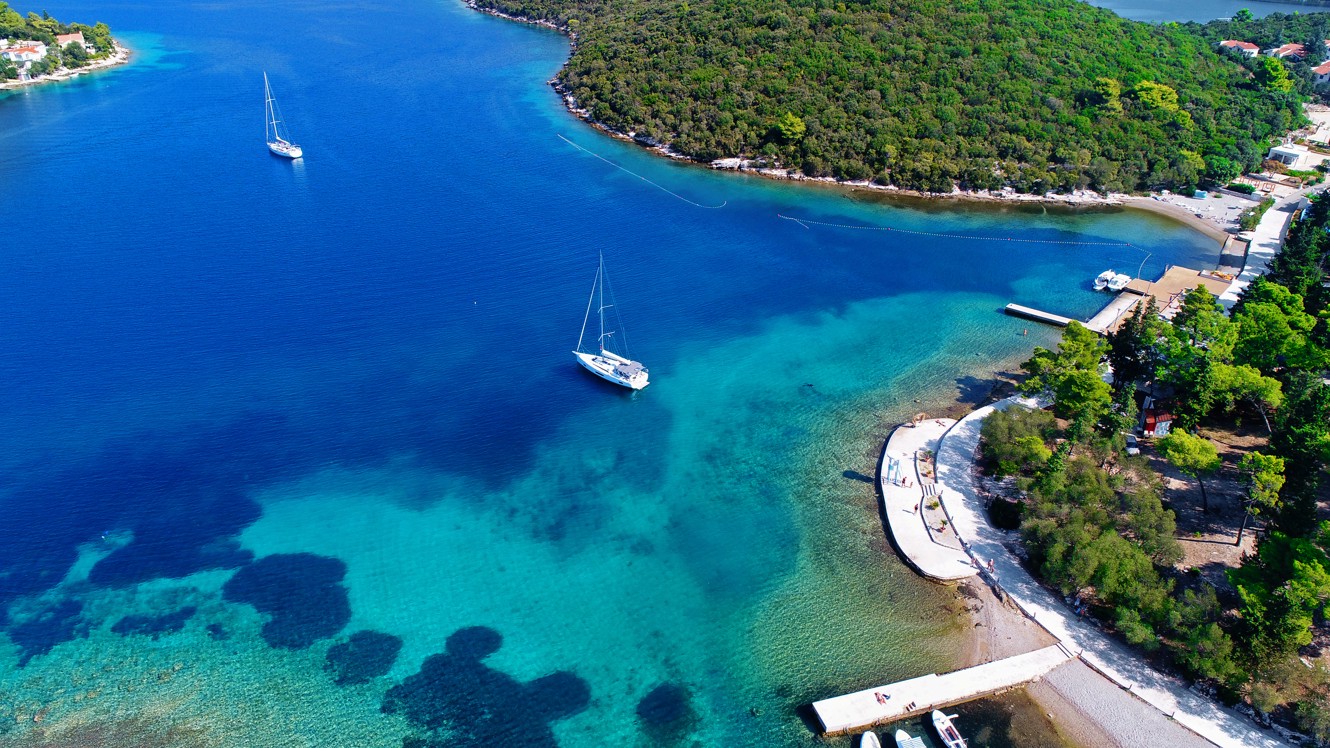 Luftaufnahme der Bucht bei Port9 Planet Camping by Aminess, Korcula, mit leuchtend türkisfarbenem Wasser, einer kleinen Insel, verankerten Booten und üppigen grünen Kiefernwäldern, die den Campingplatz umgeben, unter einem klaren blauen Himmel.
