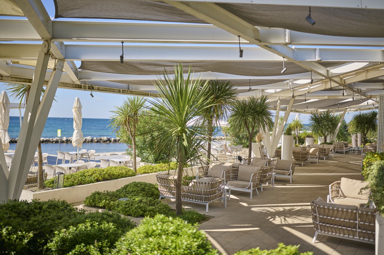 Blick auf den Strand aus dem schattigen Bereich des Restaurants im Aminess Planet Camping Maravea Resort, mit Mobilheimen und Grünflächen – ein perfekter Ort zum Entspannen und Genießen einer Mahlzeit