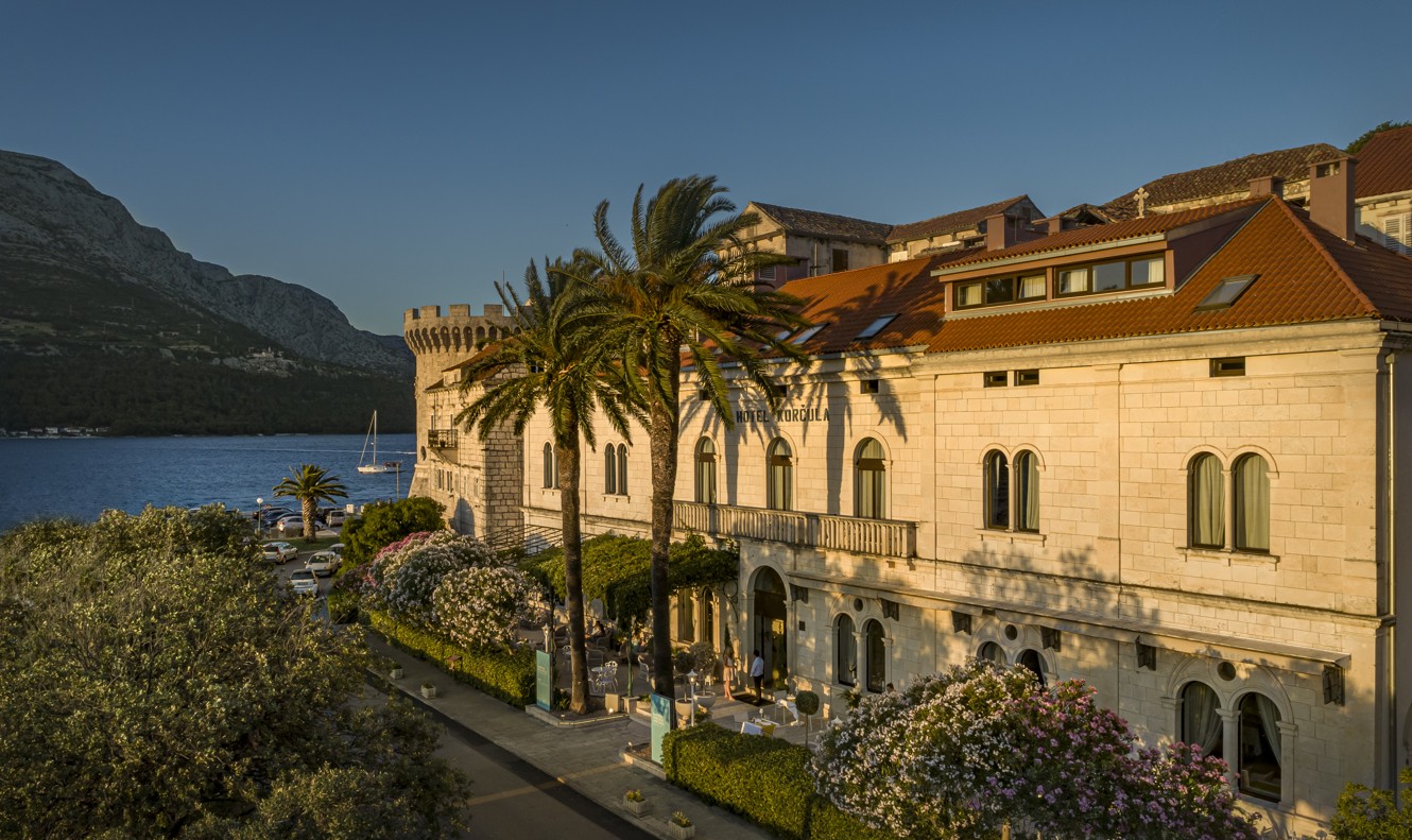 Aminess Younique Korcula Heritage Hotel, a historic building with red-tiled roofs in Korcula's Old Town near the sea, located beside Tower Kanalevic, surrounded by palm trees and blooming gardens, captured during golden hour.