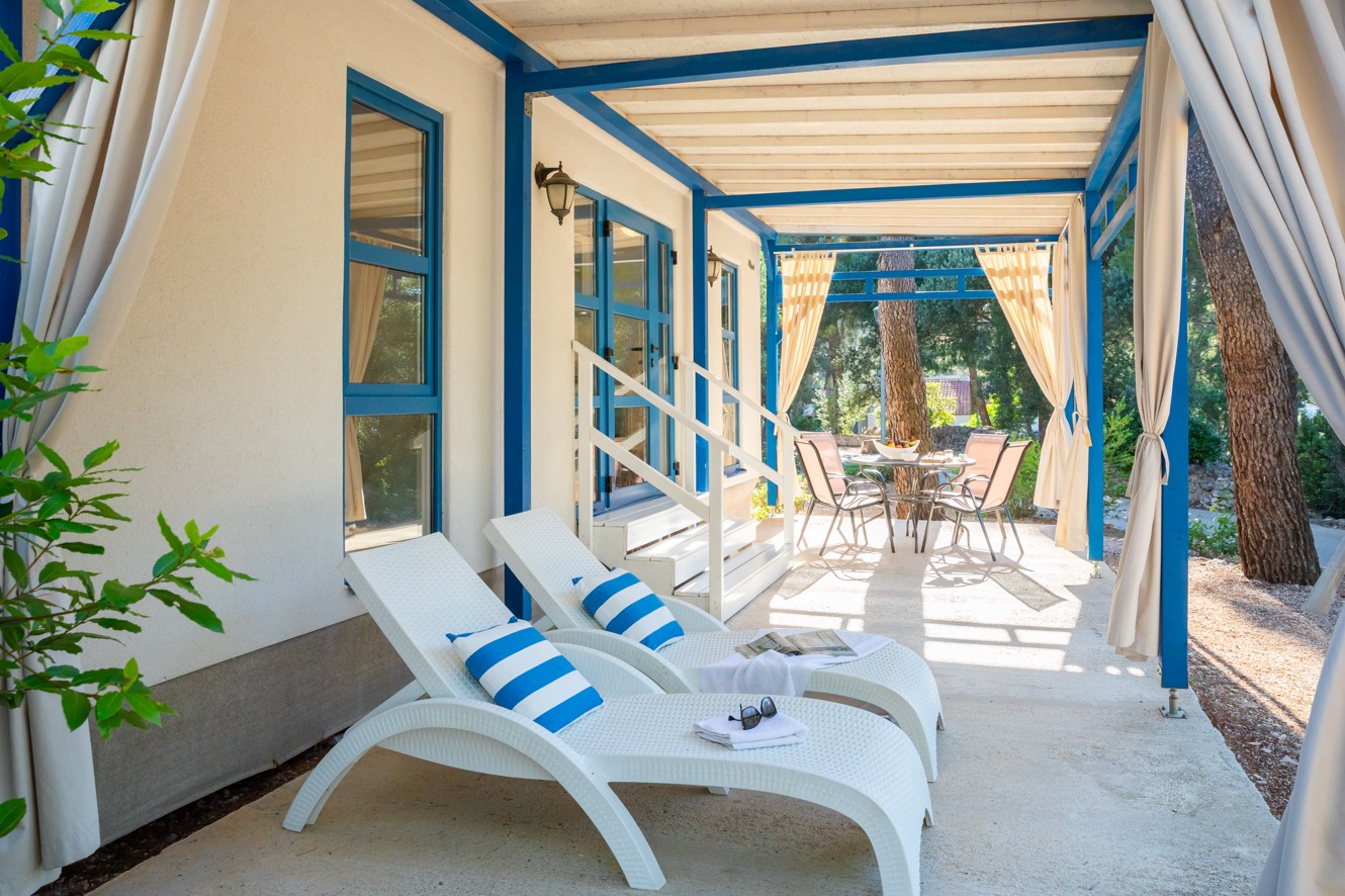 Terras voor Marko Polo Holiday Home bij Port9 Planet Camping door Aminess, Korcula, met blauw-omlijste structuren omgeven door pijnbomen onder een heldere hemel.