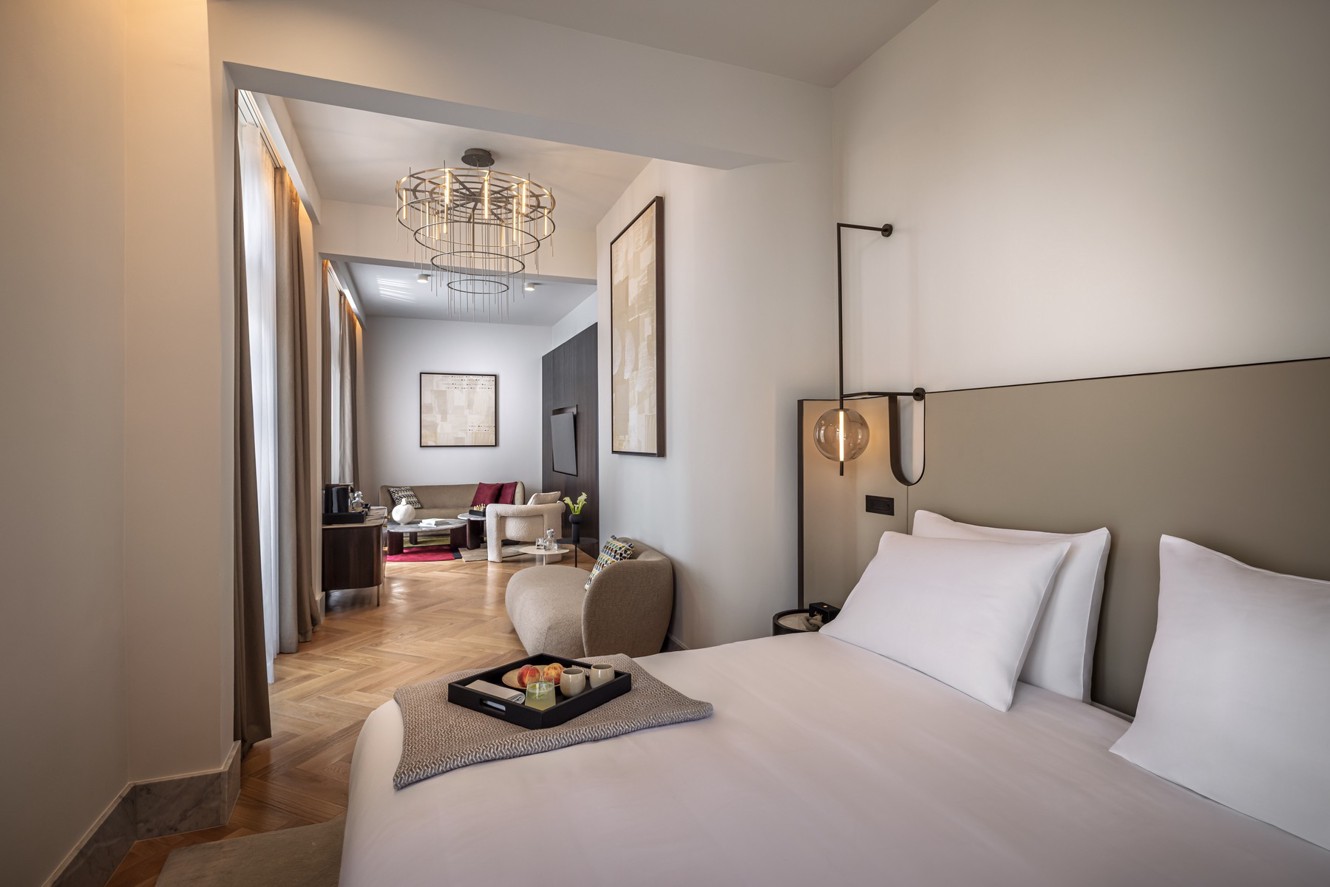 Aminess hotel suite with a bedroom, living area and breakfast on the bed.