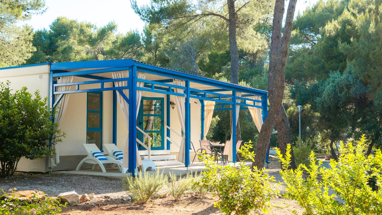 Exterieur zicht op Marko Polo Holiday Home bij Port9 Planet Camping door Aminess, Korcula, met blauw-omlijste structuren omgeven door pijnbomen onder een heldere hemel.