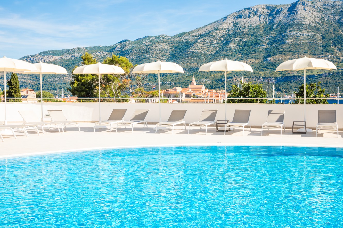 Područje vanjskog bazena u Maradiso Marko Polo by Aminess u Korčuli s ležaljkama za sunčanje, suncobranima i prekrasnim pogledom na okolna brda.