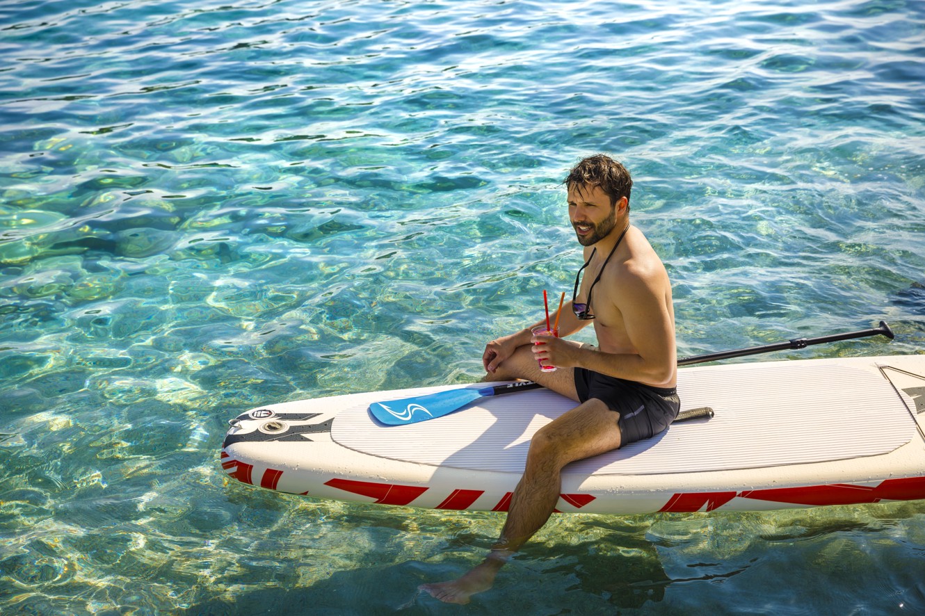 Un uomo che si diverte a fare stand-up paddleboarding nelle acque turchesi vicino ad Aminess Port9 Planet Camping Korcula, circondato da un tranquillo scenario naturale