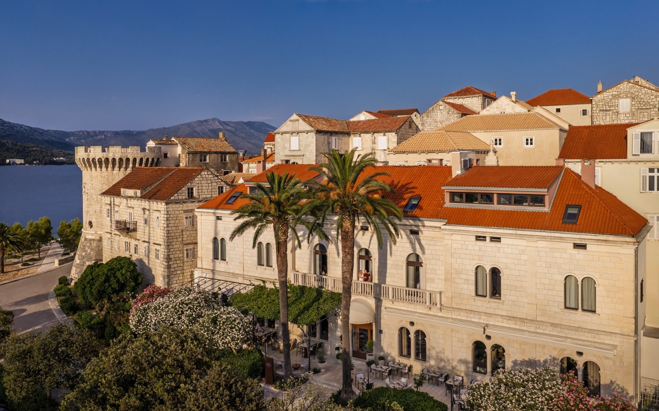 Historic stone Aminess hotel by the sea with palm trees and red roofs.