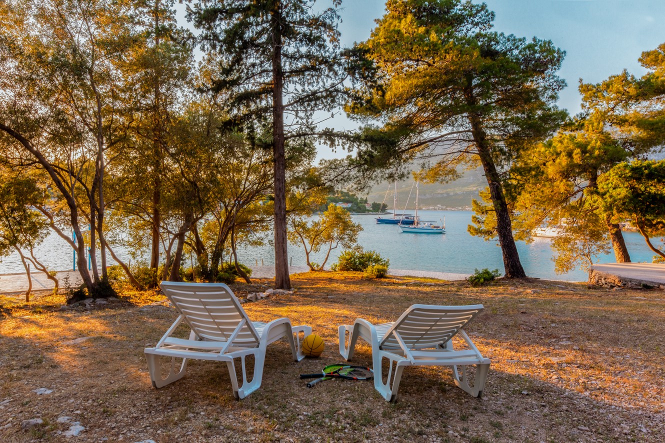 Ein malerischer Meerblick-Campingplatz bei Aminess Port9 Planet Camping, Korcula, mit Sonnenliegen unter hohen Bäumen, mit Blick auf das Wasser und umgeben von bewaldeter Landschaft