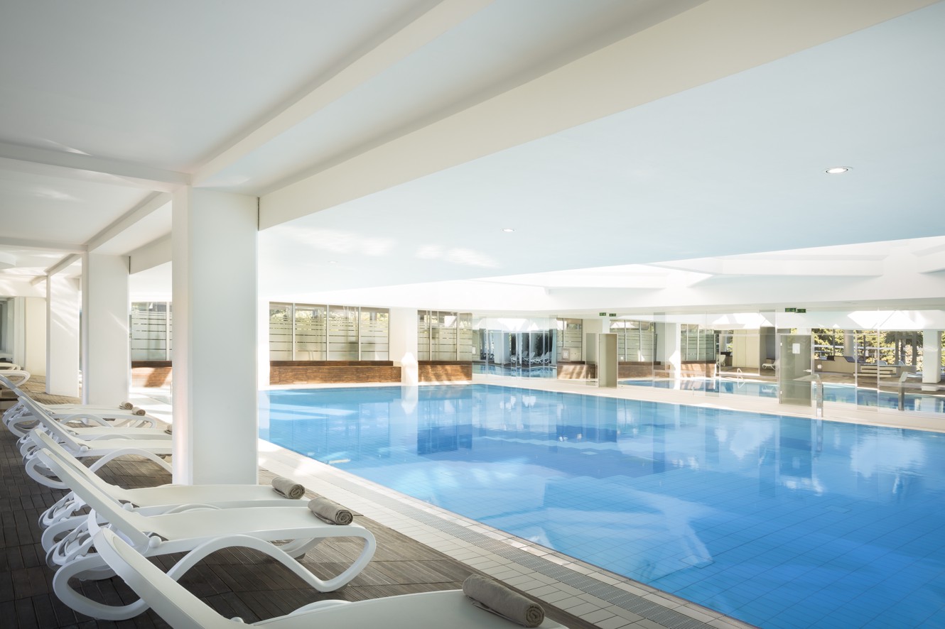 Vista della piscina interna, sedie a sdraio con asciugamani e vista del verde all'esterno.