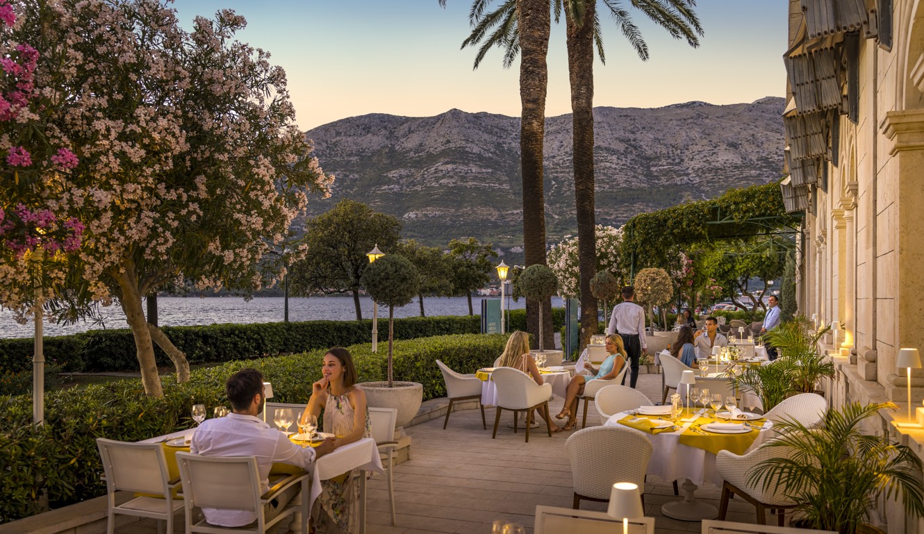 Guests enjoying a romantic dinner on the terrace of 7 Seas Restaurant & Bar at Aminess Korcula Heritage Hotel at sunset, surrounded by blooming oleander trees, palm trees, and a view of the Peljesac hills across the sparkling sea.