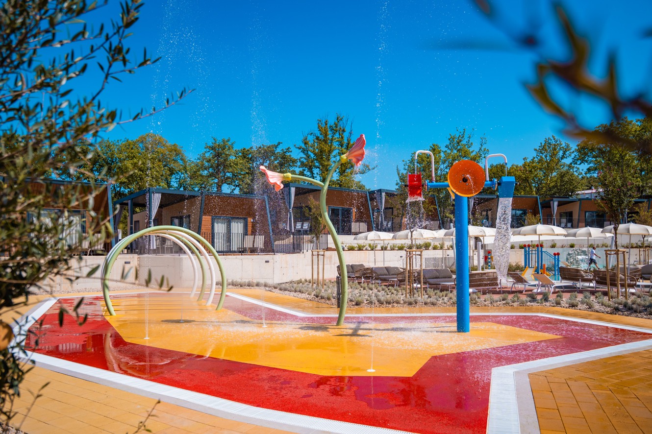 Pisano vodno igrišče z interaktivnimi elementi in pršilci, obdano z mobilnimi hiškami v Aminess Style Camping Atea Resortu.