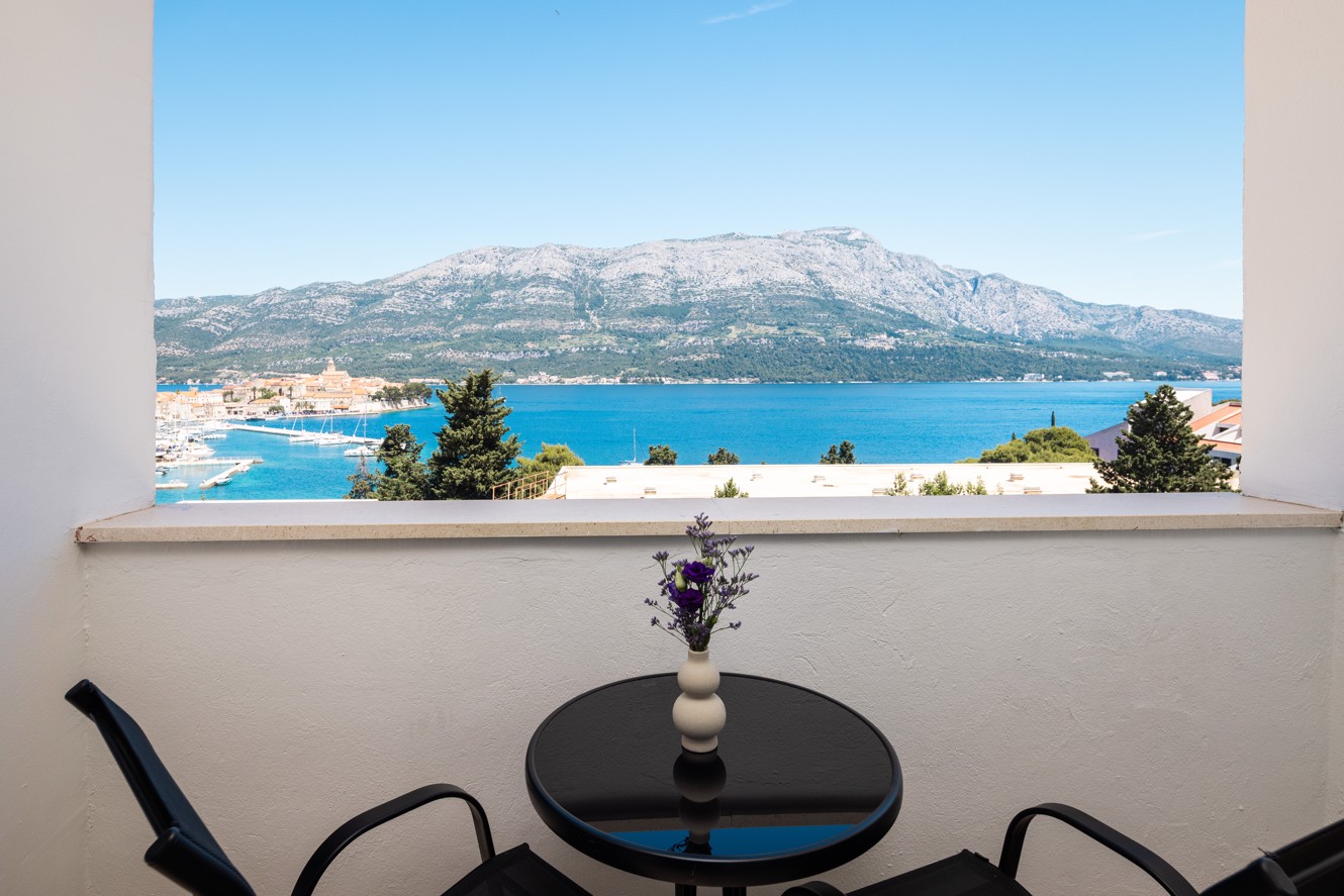 Scenski pogled iz sobe u Maradiso Marko Polo by Aminess na Korčuli, prikazuje marinu, povijesne zgrade i bujno zelenilo kroz prozor.