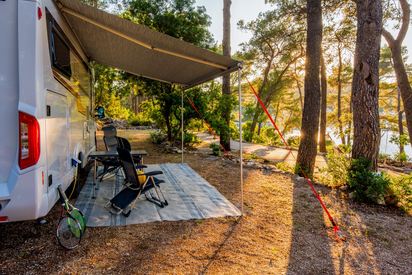 Ein Auto-Campingplatz bei Aminess Port9 Planet Camping Korcula, mit einem Wohnmobil geparkt unter Kiefern, einem schattigen Sitzbereich und Blick auf die umliegende Naturlandschaft