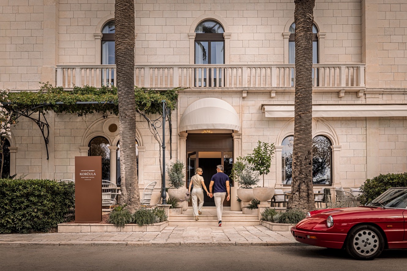 A couple enters Aminess Younique Korcula Heritage Hotel beside a red car and palm trees.