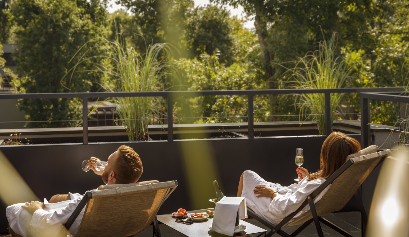 Couple relaxing on a terrace in lounge chairs, enjoying drinks with a view of lush greenery in the background.