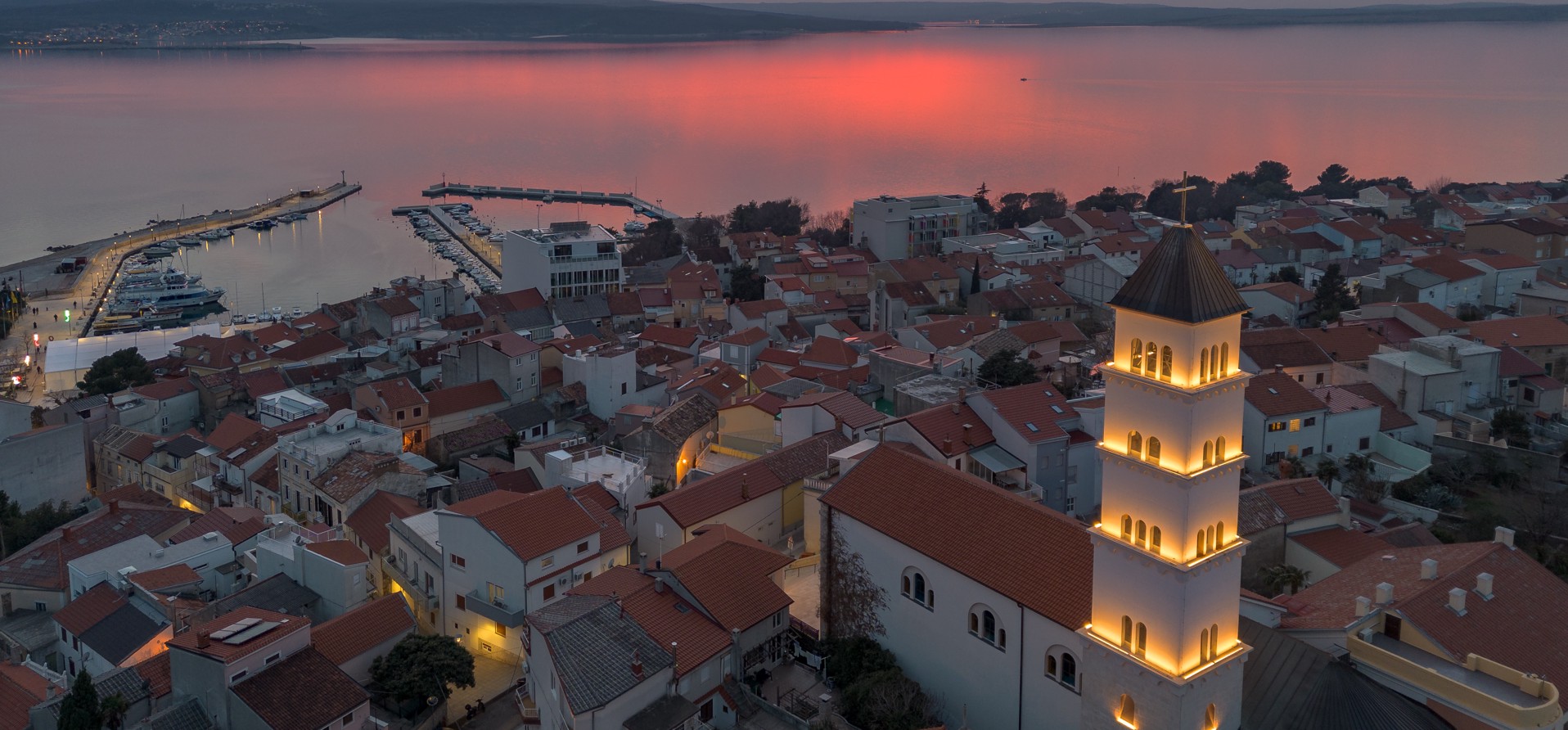 Pogled iz zraka na Crikvenicu pri sončnem zahodu, z osvetljenim zvonikom cerkve in Jadranskim morjem v ozadju.