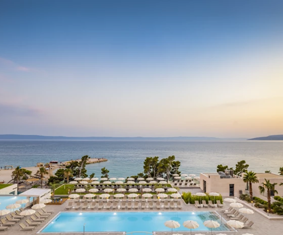 Panoramski pogled na hotelski resort s vanjskim bazenom, sunčalištem i otvorenim pogledom na more u mirnoj i elegantnoj atmosferi.