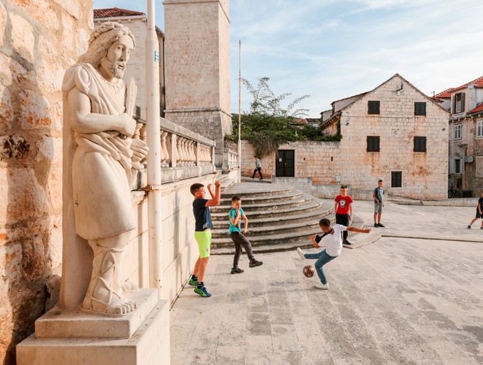 Una statua di pietra di un santo in primo piano, con bambini che giocano a calcio sullo sfondo nella piazza principale dell'Isola di Brač, Croazia, mettendo in risalto la fusione di tradizione e vita locale.