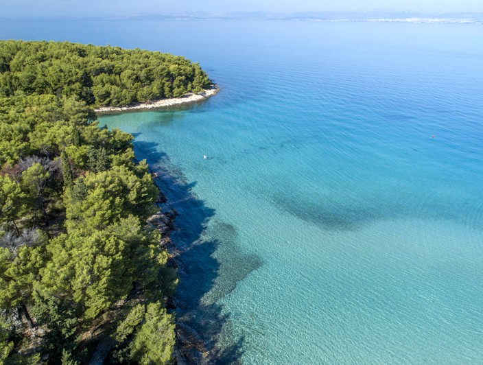 Pogled iz zraka na stjenovitu plažu na otoku Braču, Hrvatska, okruženu bujnom zelenom vegetacijom i kristalno plavim Jadranskim morem.